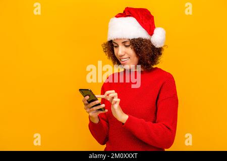 Giovane donna in cappello di babbo natale che tiene il telefono cellulare, leggendo il messaggio di vacanza, si congratula con gli amici felice anno nuovo Foto Stock