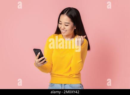 Giovane donna asiatica eccitato ha ricevuto un buon messaggio sul telefono cellulare, felice con grande notizia in piedi su sfondo rosa Foto Stock
