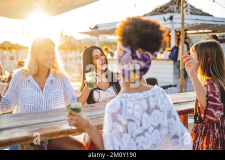 quattro donne che si divertono al bar sulla spiaggia, giovani amiche che ridono e chiacchierano, bevono un drink e trascorrono del tempo in vacanza, in vacanza e in estate Foto Stock