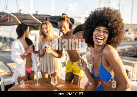Bella giovane afroamericana sorridente e danzante guardando la macchina fotografica, gruppo di amici rilassarsi su yacht di lusso e bere champagne, ha Foto Stock