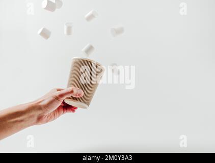 Tazza di carta con bevanda calda, marshmallows volanti in mano maschile. Sfondo bianco. Caffè, latte, cappuccino, cacao. Tazza di carta monouso. Riciclaggio ed e Foto Stock