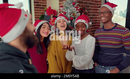 Gruppo multietnico di dipendenti che registra video in ufficio durante le feste, riprese con la fotocamera durante la festa della vigilia di natale. Gente allegra che scatta foto e che si diverte in vacanza di Natale. Foto Stock