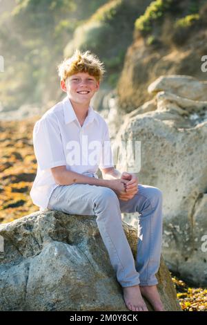 Tween Boy siede su Una roccia alla spiaggia durante l'ora d'oro Foto Stock