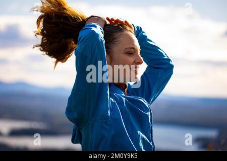 Una donna in una giacca blu fissa i suoi capelli che si affaccia su un lago Foto Stock