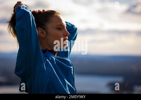 Una donna in una giacca blu fissa i suoi capelli che si affaccia su un lago Foto Stock