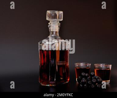 Liquore di chokeberry fatto in casa su sfondo nero, una bottiglia, due bicchieri con una bevanda e un mazzo di bacche da giardino mature. Primo piano. Foto Stock