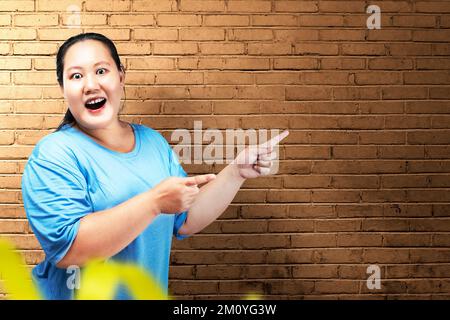 Asian grasso donna sovrappeso con un'espressione eccitata che indica qualcosa con uno sfondo muro di mattoni Foto Stock