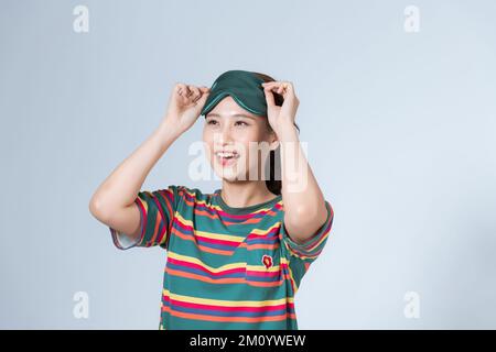 Giovane donna eccitata ridendo tenendo maschera di sonno sulla fronte e guardando da sotto di esso, divertendosi sulla festa pajama, isolato su sfondo bianco Foto Stock