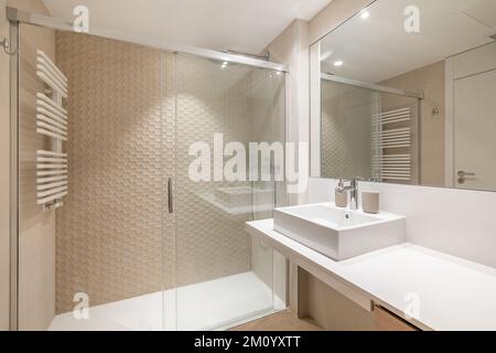 Interni spaziosi di un moderno bagno piastrellato con lavabo bianco, grande specchio e zona doccia. Foto Stock