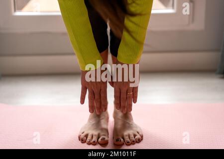 Alto angolo di raccolto irriconoscibile donna barefoted in piedi sul tappeto e piegarsi in avanti mentre fare esercizio stretching durante la sessione di yoga in appartamento leggero Foto Stock