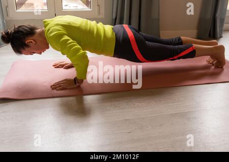 Vista laterale di determinata giovane femmina con i capelli scuri in activewear che fa la posa di Chaturanga Dandasana mentre pratica lo yoga sul tappetino in appartamento Foto Stock