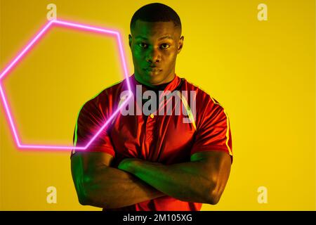 Neon esagonale troppo fiducioso giovane sportivo afro-americano con le braccia incrociate su sfondo giallo. Composito digitale, sport, atleta, fitness, copia Foto Stock
