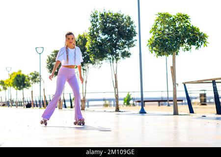 Ritratto di bella ragazza bionda adolescente con cool pigtail, maglietta bianca e pantaloni rosa flared pattinaggio su rulli nel parco Foto Stock