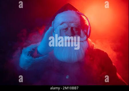 Babbo natale ascolta musica con le cuffie durante una festa. Luce al neon blu rossa e fumo. Foto Stock