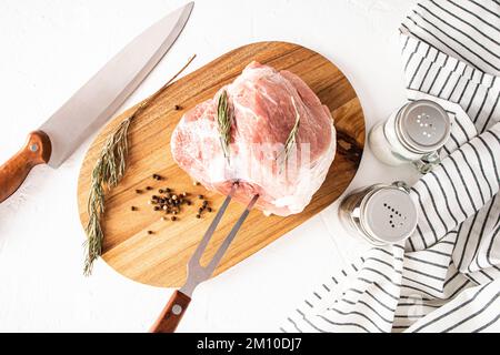 Un grosso pezzo di prosciutto di maiale si trova su un tagliere di rosmarino per preparare un piatto da menu. vista dall'alto. il processo di preparazione del taglio della carne Foto Stock