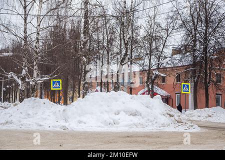 Una grande nevicata bianca sullo sfondo di una strada cittadina con alberi. Sulla strada si trova la neve bianca in alto mucchi. Paesaggio urbano invernale. Nuvoloso giorno d'inverno, luce soffusa. Foto Stock