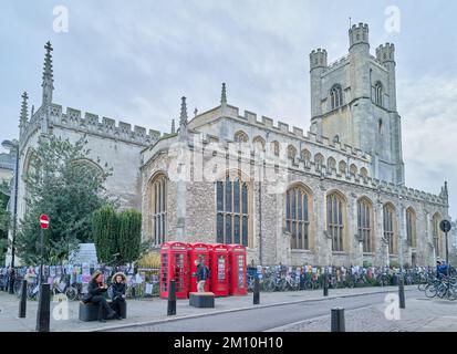 Un paio di turisti si siedono su una panchina vicino a quattro cabine telefoniche rosse fuori dalla chiesa di St Mary, la chiesa universitaria di Cambridge, Inghilterra. Foto Stock