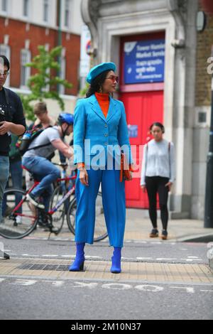 Elegante donna in berretto blu, stivali blu, pantaloni blu, giacca blu, dolcevita arancione, Occhiali da sole e una pochette arancione su una strada londinese Foto Stock
