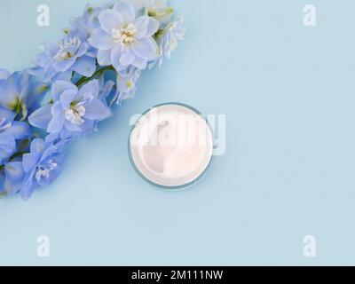 Crema idratante in vaso di vetro aperto e fiore blu di delfinio su sfondo blu. Bianco per bottiglie cosmetiche con acido ialuronico. Mockup, copia spazio, in alto Foto Stock