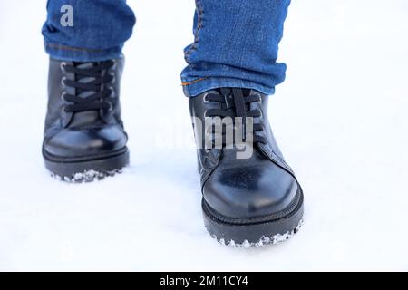 Gambe femminili in pelle nera stivali con lacci e jeans sulla neve. Donna che cammina sulla strada d'inverno, scarpe calde per il freddo Foto Stock