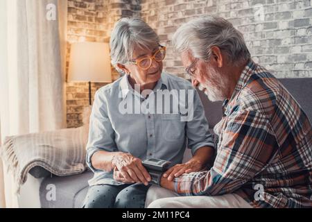 Primo piano e ritratto di vecchia donna che controlla la pressione del marito a casa seduto non lui divano. Un uomo maturo che si sente male e malato misurando l'ipertensione con una macchina. Foto Stock