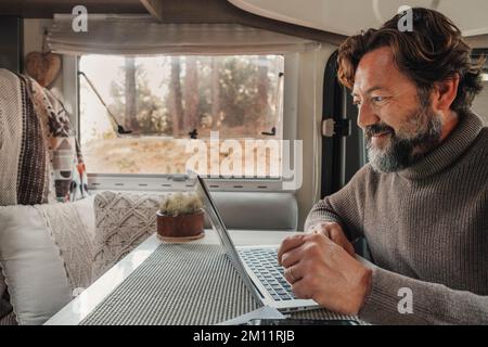 Un uomo lavora su un computer portatile all'interno di un camper seduto al tavolo della scrivania. Stile di vita moderno del lavoro di connessione online. Persone e nomade digitale van vita lifestyle. Natura all'aperto fuori dalla finestra. Libertà Foto Stock