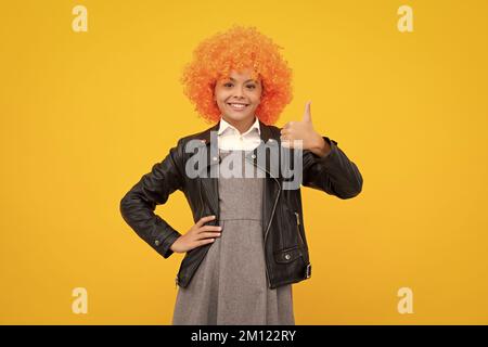 Festa di compleanno per bambini. Capretto divertente in parrucca clown riccio isolato su sfondo giallo. Faccia felice della ragazza, emozioni positive e sorridenti. Foto Stock