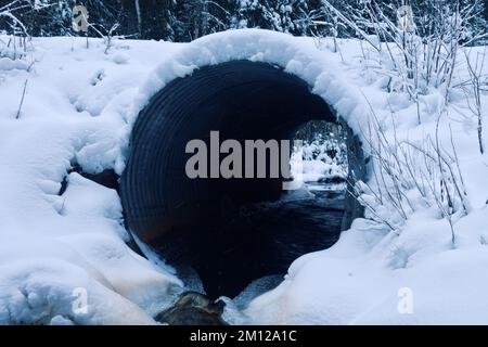 Il canale sotto la strada forestale è coperto di neve in inverno, ma lo scarico d'acqua continua, ghiaccio - ruscello libero Foto Stock