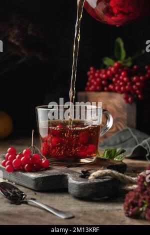 Il tè di viburnum è versato in una tazza da una teiera di vetro, processo di versamento. Autunno stagionale bevanda sana. Orientamento verticale Foto Stock