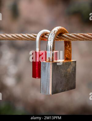 Primo piano di due lucchetti d'amore arrugginiti fissati su una corda d'acciaio con messa a fuoco selettiva e fondo bokeh. Immagine a toni con colori desaturati Foto Stock