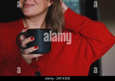 primo piano di una donna felice che tiene una tazza grigia di caffè, indossando un caldo maglione rosso accogliente Foto Stock