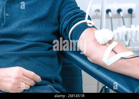 Tema della donazione. L'uomo dona sangue in ospedale. La mano della persona con l'ago e IV. Primo piano. Il donatore si siede sulla sedia. Sfondo.. Foto Stock