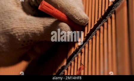 Primo piano delle mani dell'uomo in guanti di protezione martellando un perno nella parete. Concetto di riparazione e ristrutturazione, utensili professionali e per lavoratori. Foto Stock