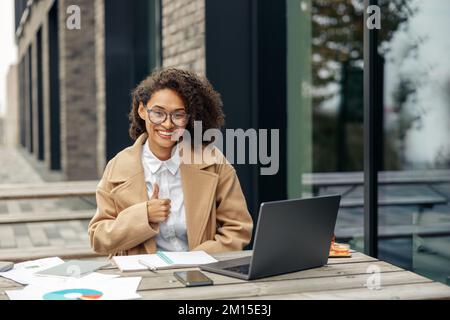 Donna africana sorridente che mostra il pollice in su mentre lavora il laptop fuori vicino all'edificio dell'ufficio Foto Stock