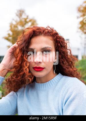Deliziati le giovani donne millennial con lunghi capelli ricci di zenzero e labbra rosse in abiti casual guardando la macchina fotografica mentre si è in piedi nel parco cittadino Foto Stock