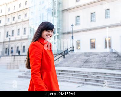 Allegra donna adulta con lunghi capelli scuri in cappotto rosso brillante che cammina vicino alle scale del vecchio edificio in strada di Madrid mentre guarda la macchina fotografica Foto Stock