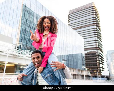 Allegro giovane etnico maschile millenario in abiti casual sorridendo felicemente, mentre dando giro a spalla a ragazza felice mostrando due dita gesto Foto Stock