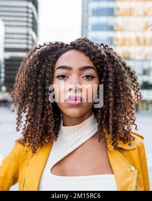 Modello femminile afro-americano fiducioso con capelli ricci scuri e piercing al naso in elegante giacca in pelle gialla in piedi sulla strada vicino moderno b Foto Stock