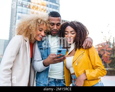 Allegro giovane multirazziale Signore in abiti eleganti sorridendo felicemente e abbracciando positivo maschio migliore amico utilizzando il telefono cellulare sulla piazza della città Foto Stock