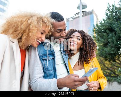 Allegro giovane multirazziale Signore in abiti eleganti sorridendo felicemente e abbracciando positivo maschio migliore amico utilizzando il telefono cellulare sulla piazza della città Foto Stock