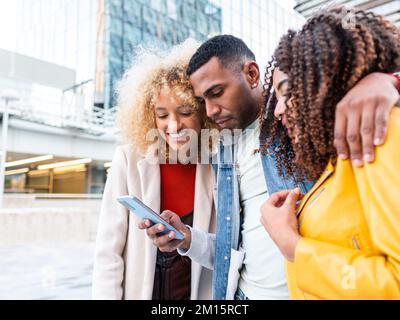 Allegro giovane multirazziale Signore in abiti eleganti sorridendo felicemente e abbracciando positivo maschio migliore amico utilizzando il telefono cellulare sulla piazza della città Foto Stock