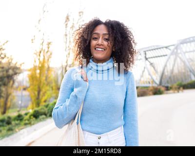 Allegra giovane ragazza etnica con acconciatura afro in dolcevita blu in piedi sulla strada della città e tenendo borsa a corda mentre si guarda la macchina fotografica Foto Stock