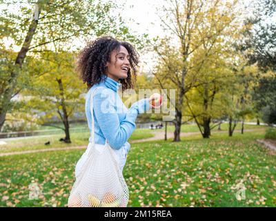 Vista laterale di una donna ispanica sorridente in abiti casual con borsa a corda che cammina lungo la strada del parco con mela matura il giorno d'autunno Foto Stock