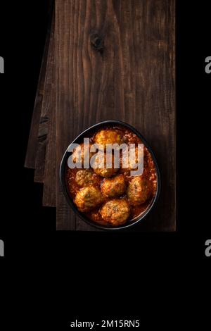 Vista dall'alto della ciotola nera con deliziose polpette in salsa di pomodoro con erbe poste su un mucchio di tavole di legno nella stanza scura Foto Stock