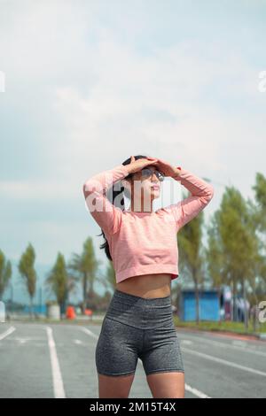 Sportivo femminile in abbigliamento sportivo e occhiali in piedi guardando lontano sulla pista di corsa preparandosi per l'allenamento allo stadio Foto Stock