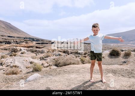 Corpo pieno di capretto a piedi nudi sorridente in abiti estivi in piedi con braccia distese su terreno sabbioso nella valle del deserto contro montagne e loo Foto Stock