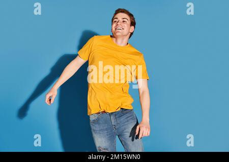 Giovane uomo energico in t-shirt gialla e jeans che guarda lontano e sorridente mentre salta su sfondo blu Foto Stock