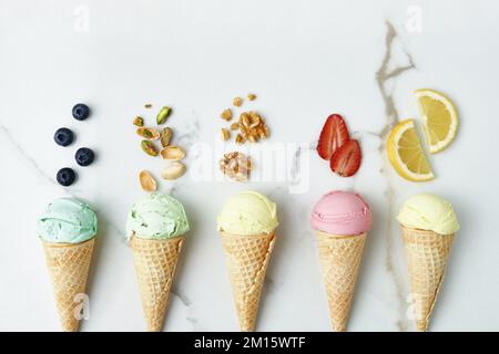 Vista dall'alto di assortiti e appetitosi coni di gelato con noci al pistacchio di mirtillo fragola e limone composto su tavola di marmo con ingredienti in kit Foto Stock