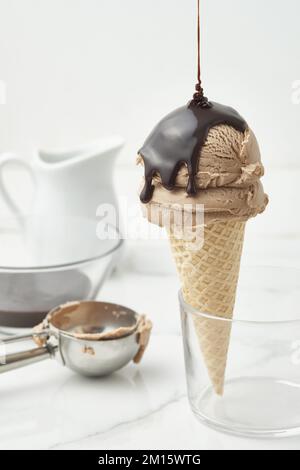 Ganache al cioccolato dolce versate su un appetitoso scoop di gelato in croccante cialde cono posto in una ciotola di vetro su un tavolo di marmo in una cucina leggera Foto Stock