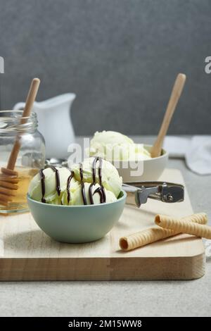 Scoop gelato yummy in ciotola con salsa di cioccolato e servito su tagliere di legno con vaso di miele e panini di waffle in cucina Foto Stock